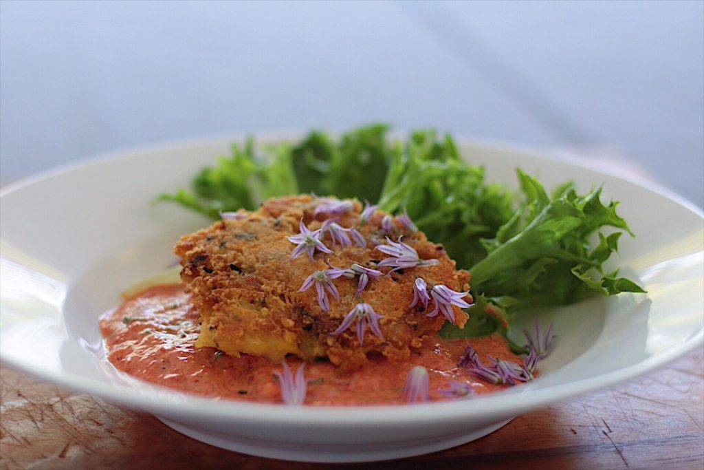 Crab cake on top of red pepper sauce garnished with lettuce leaf in a white bowl.