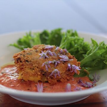 Crab cake on top of red pepper sauce garnished with lettuce leaf in a white bowl.