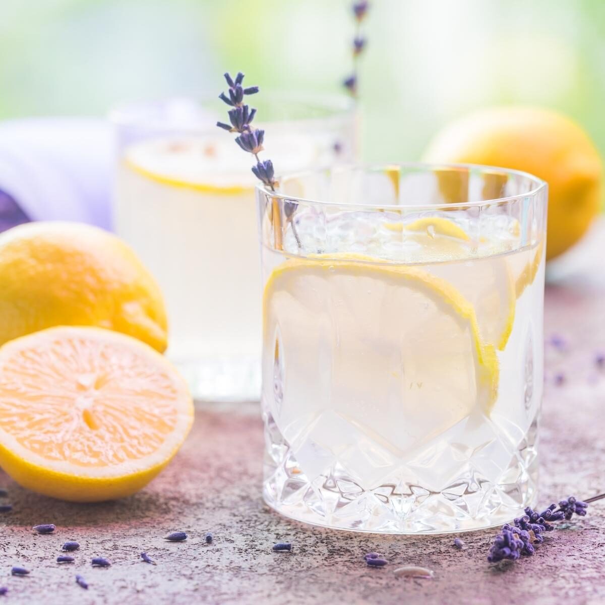 Pale yellow and lavender drink in a whisky glass. It is garnished with lemon wheel and lavender sprig with cut lemons beside on a wooden table.