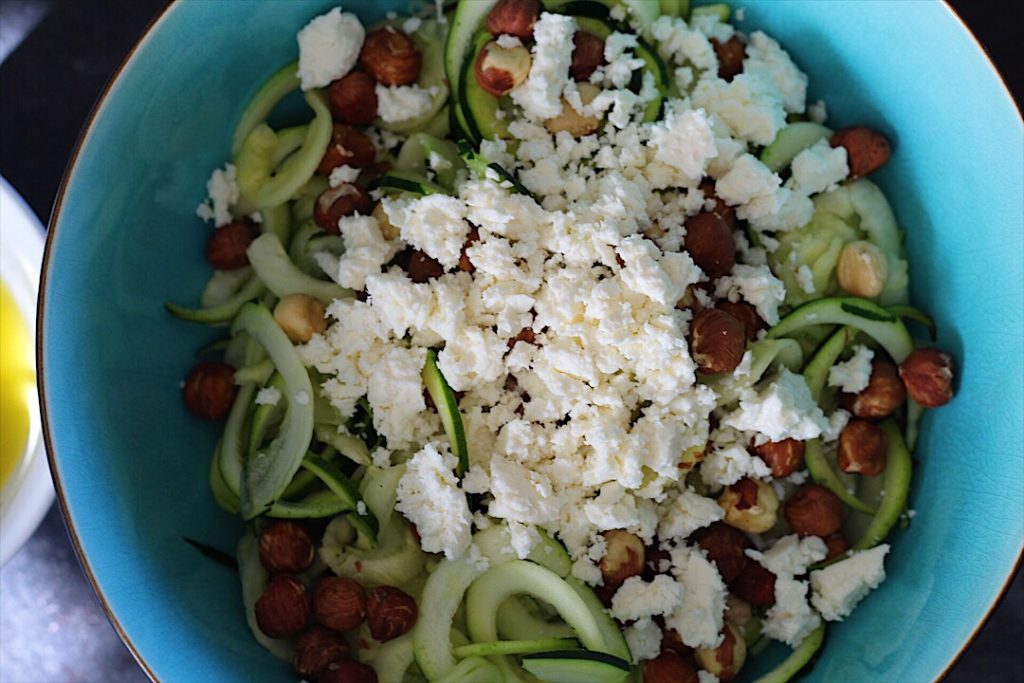 Hazelnuts and feta have been added to the top of the zucchini ribbons in the blue bowl.