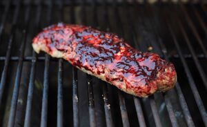 A whole pork tenderloin with deep red purple sauce on the black grates of a grill. There are some charred spots on the top.