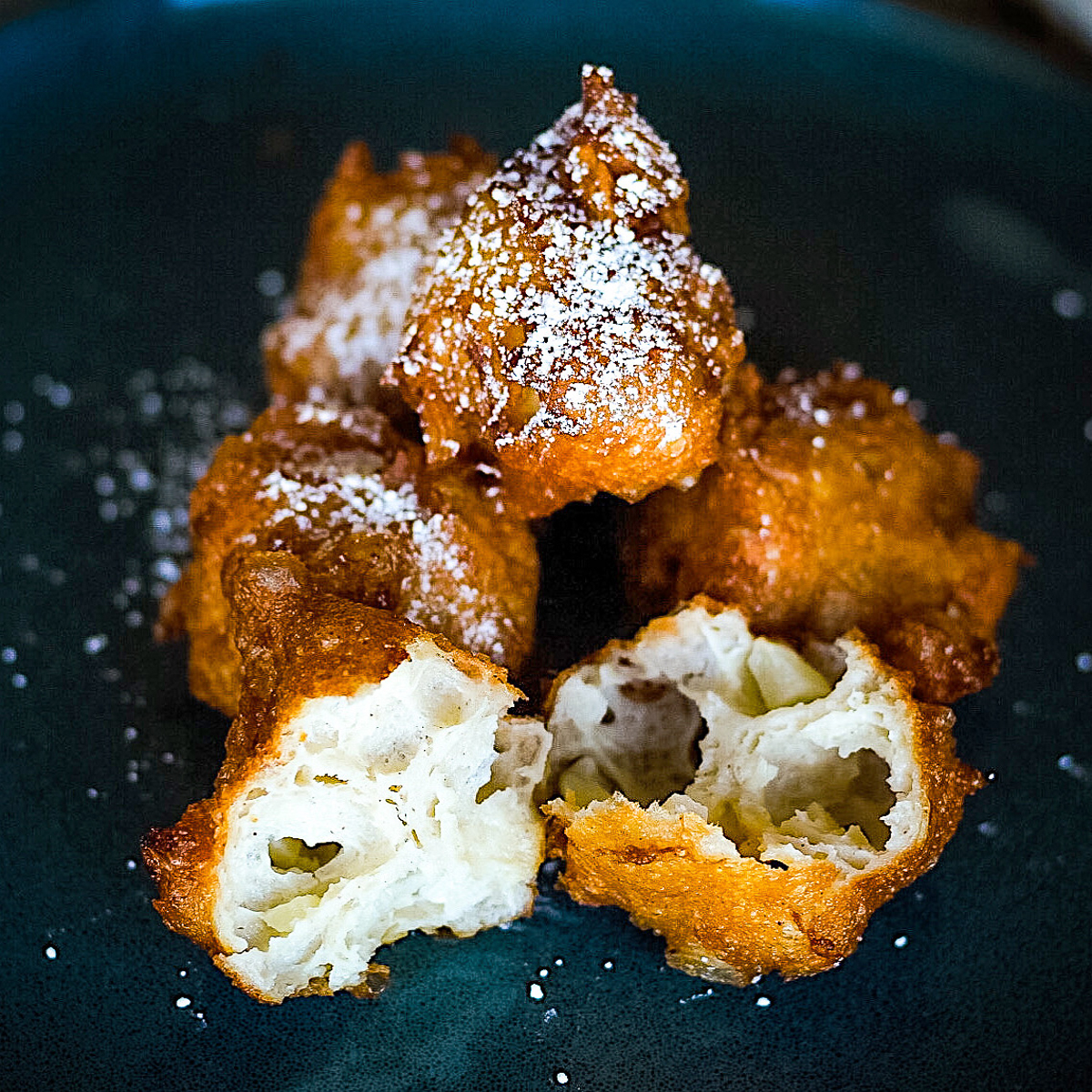 A pile of apple beignets on a dark plate, dusted with icing sugar. One fritter is broken open to show the light, airy inside of the donut.