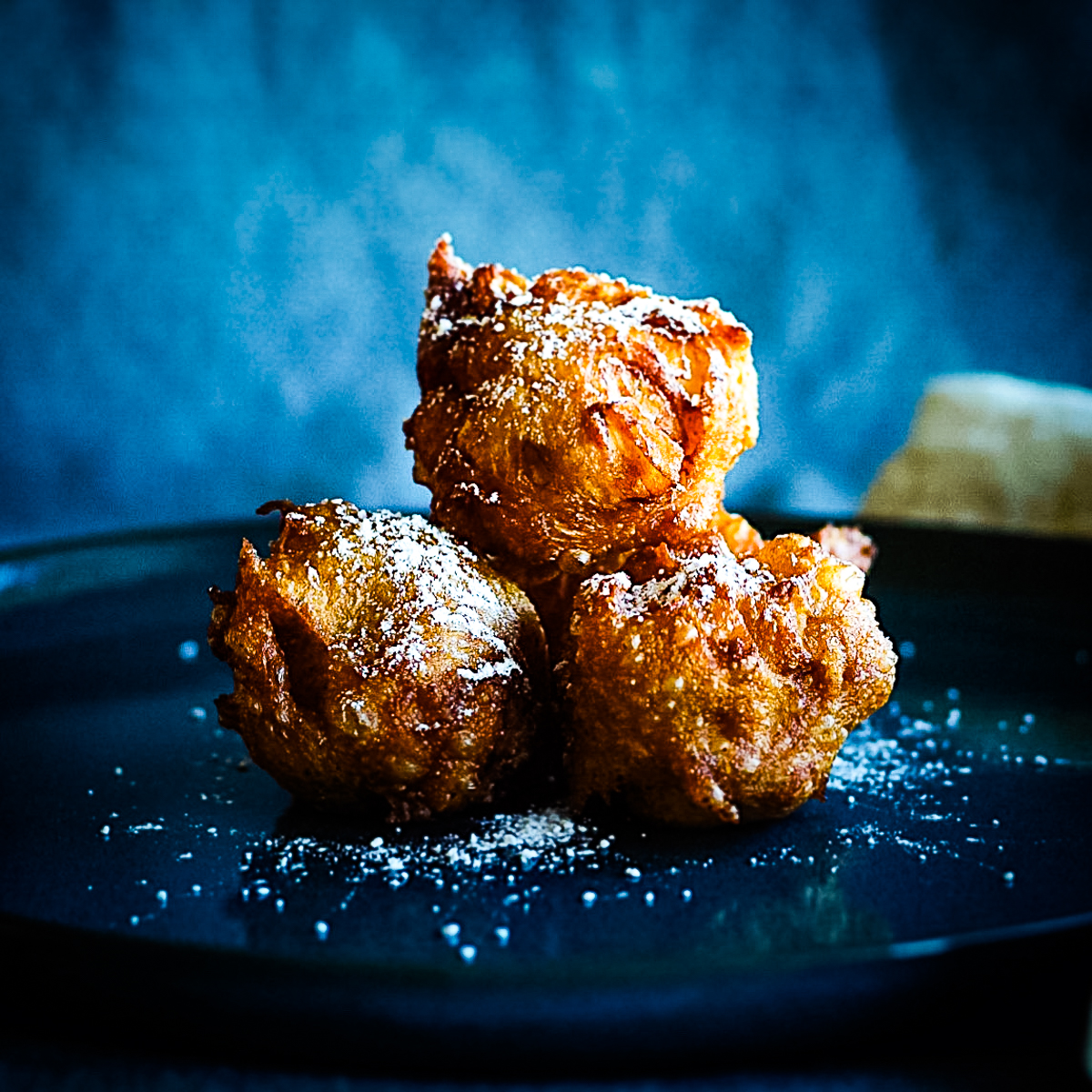 Three rustic but round apple beignets on a large round blue plate. They are golden brown and dusted with icing sugar.
