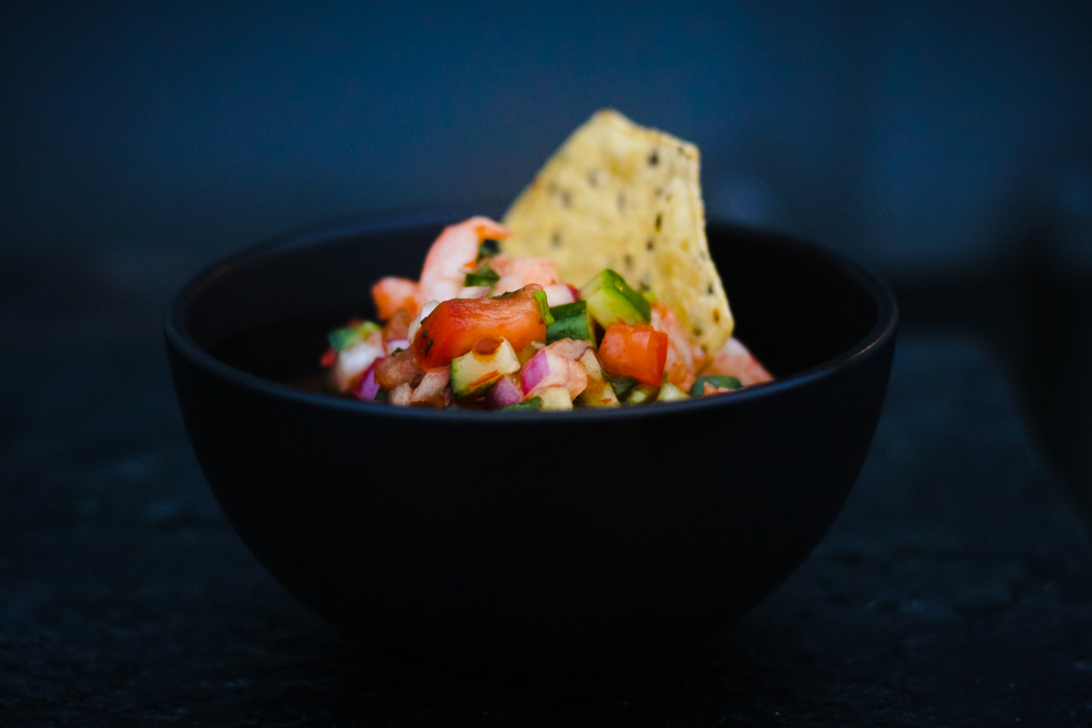 Dark bowl and background with brightly lit shrimp cocktail in center with a tortilla chip dipped into the center.