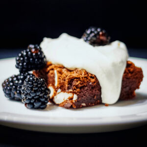 One square serving of chocolate almond brownies on a white plate with whipped cream on top and blackberries beside.