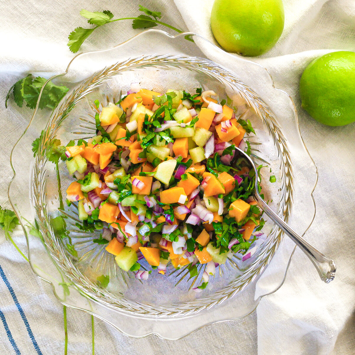 Orange, green and yellow salsa in a glass bowl with a spoon in it is on a white tea towel with limes and cilantro scattered around.