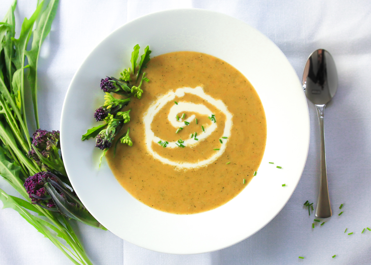 Orange broccoli cheddar soup in white bowl with a swirl of cream and garnished with green and purple broccoli florets.