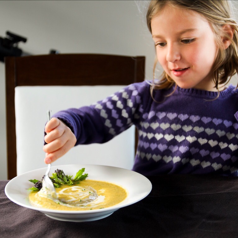 Child in purple sweater with a spoon in white bowl of broccoli soup that has a swirl of cream in it. 