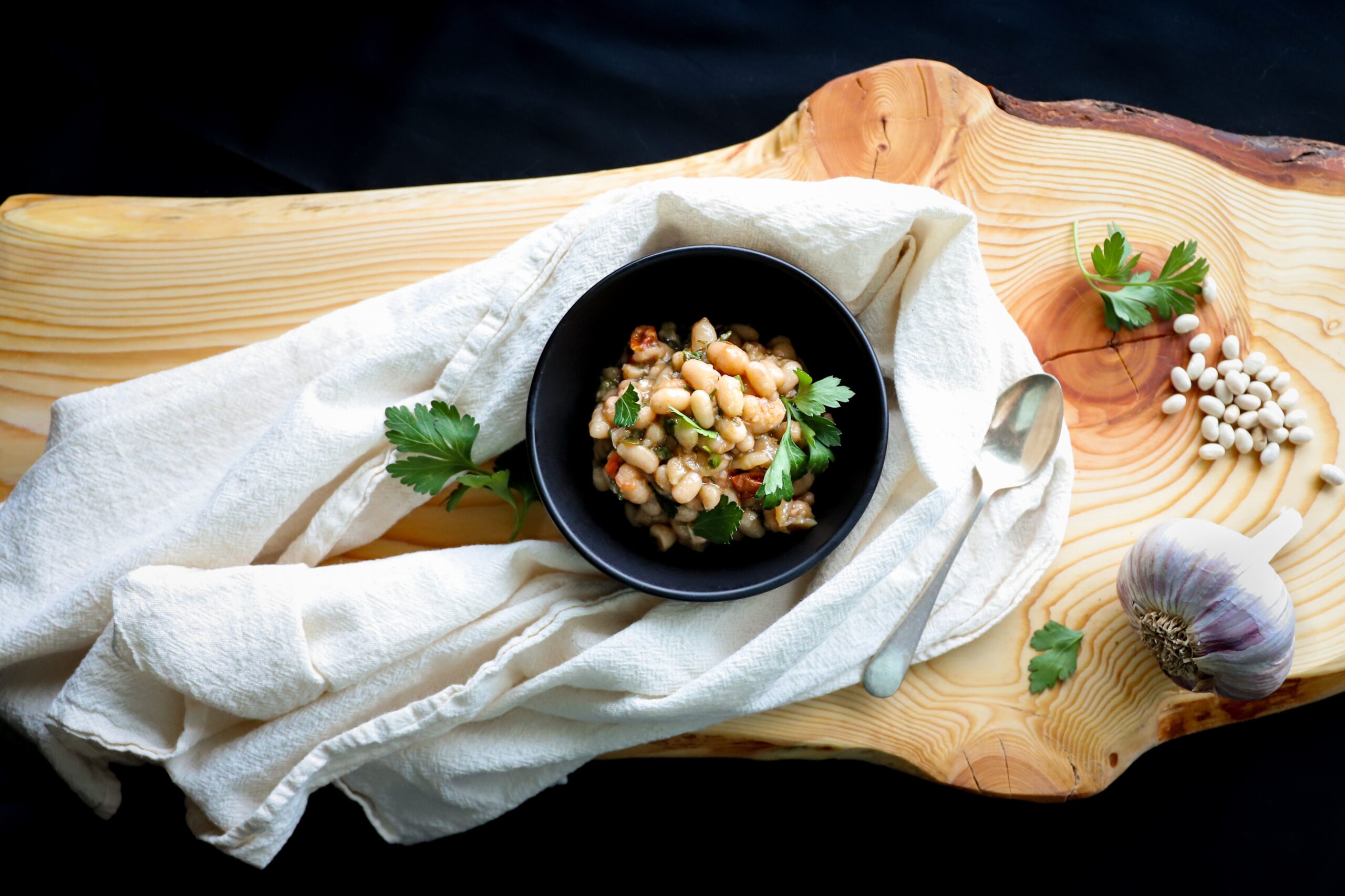 White beans in a black bowl garnished with Italian parsley. It is on a rustic wooden cutting board with a linen tea towel around the bowl. 