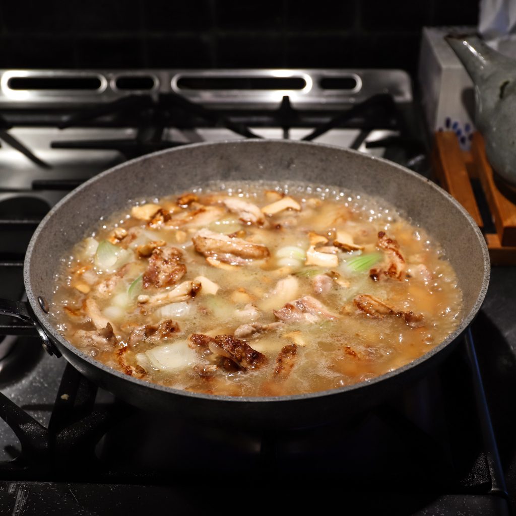 Chicken broth in a pan with chunks of browned chicken and onion is simmering with bubbles at the edges.