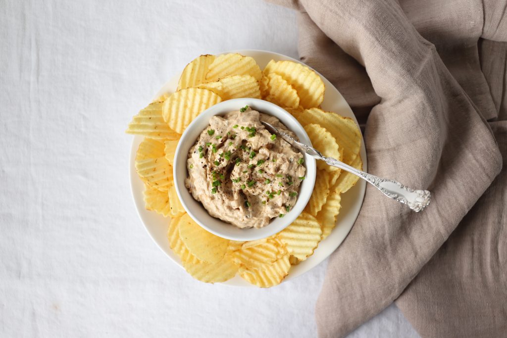 A bowl of light brown onion dip surrounded by a plate of ripple potato chips on a white tablecloth with a taupe tea towel beside.