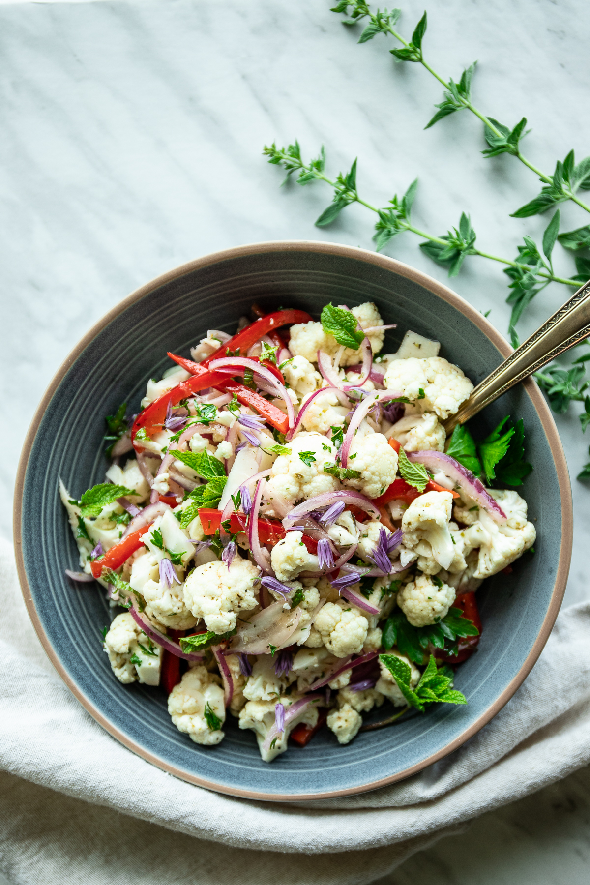 Bowl of cauliflower and red pepper salad in a stoneware bowl on a linen tablecloth with sprigs of fresh oregano around.