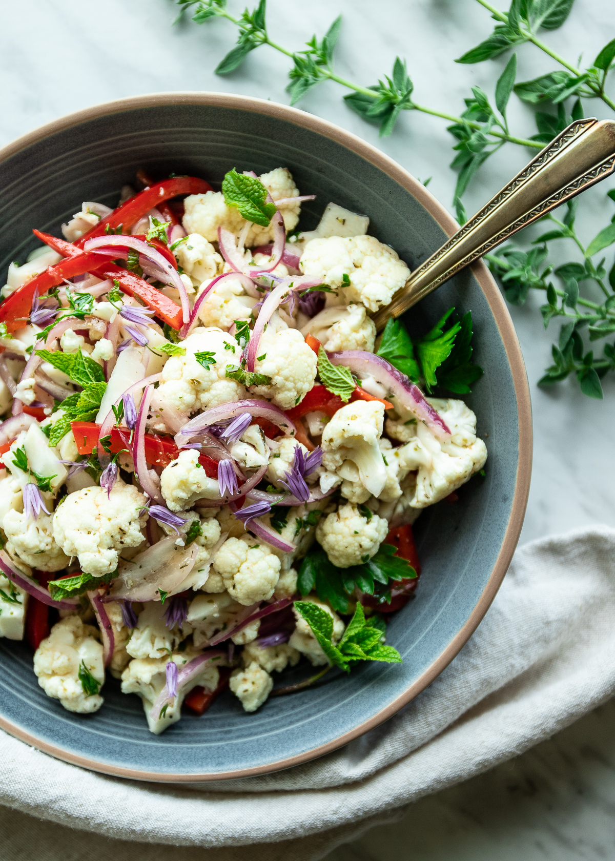 Bowl of chunky cauliflower salad in a stoneware bowl on a linen tablecloth with sprigs of fresh oregano around.