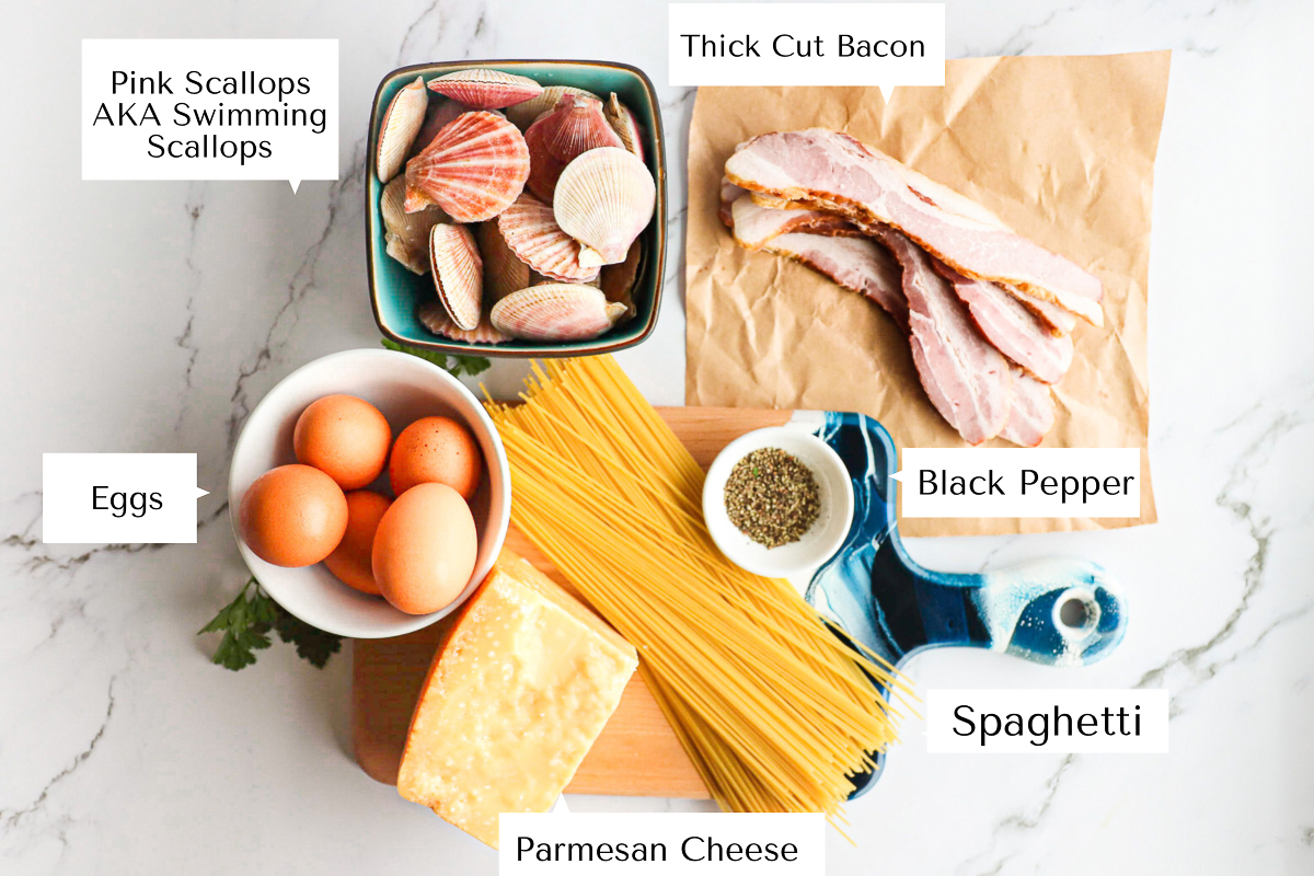 All the ingredients for the scallop and bacon pasta labelled on a cutting board including eggs, spaghetti, bacon, parmesan, pepper and scallops.