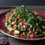 Tomato cucumber and walnut salad on a red plate and topped with lots of chopped parsley