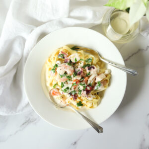 White wide rimmed bowl with creamy seafood fettuccini in it. It is on a marble countertop with a white linen napkin and a glass of white wine beside. It is an overhead shot-a flat lay.