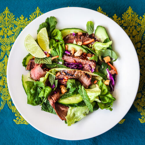 Flank Steak salad with Thai salad dressing in a white bowl set on a blue and green ornately patterned tablecloth