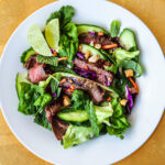 Sliced flank steak with greens and herbs in a white bowl on a gold charger