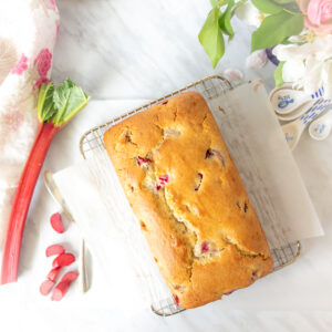 White marble countertop with a loaf of strawberry rhubarb snack cake on a cooling rack on it. Also a stalk of rhubarb, a white and pink tea towel and some white and blue measuring spoons around the perimeter of the picture.