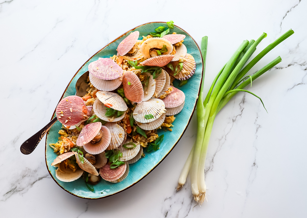 Bright pink shelled small swimming scallops in fried rice on a blue plate on a marble countertop with green onions on it