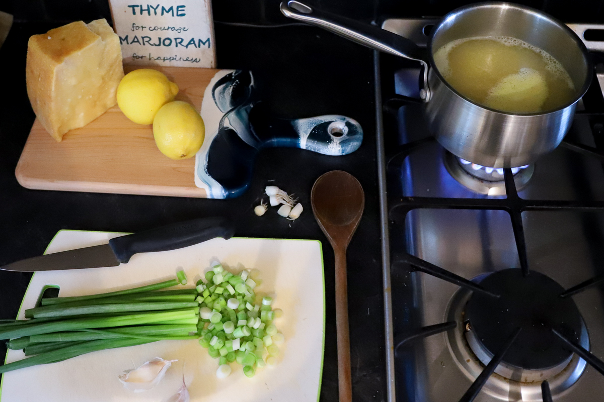 Pot of hot stock on the stove for the risotto, green onions are on the cutting board beside half chopped. Parmesan and lemons in the background.