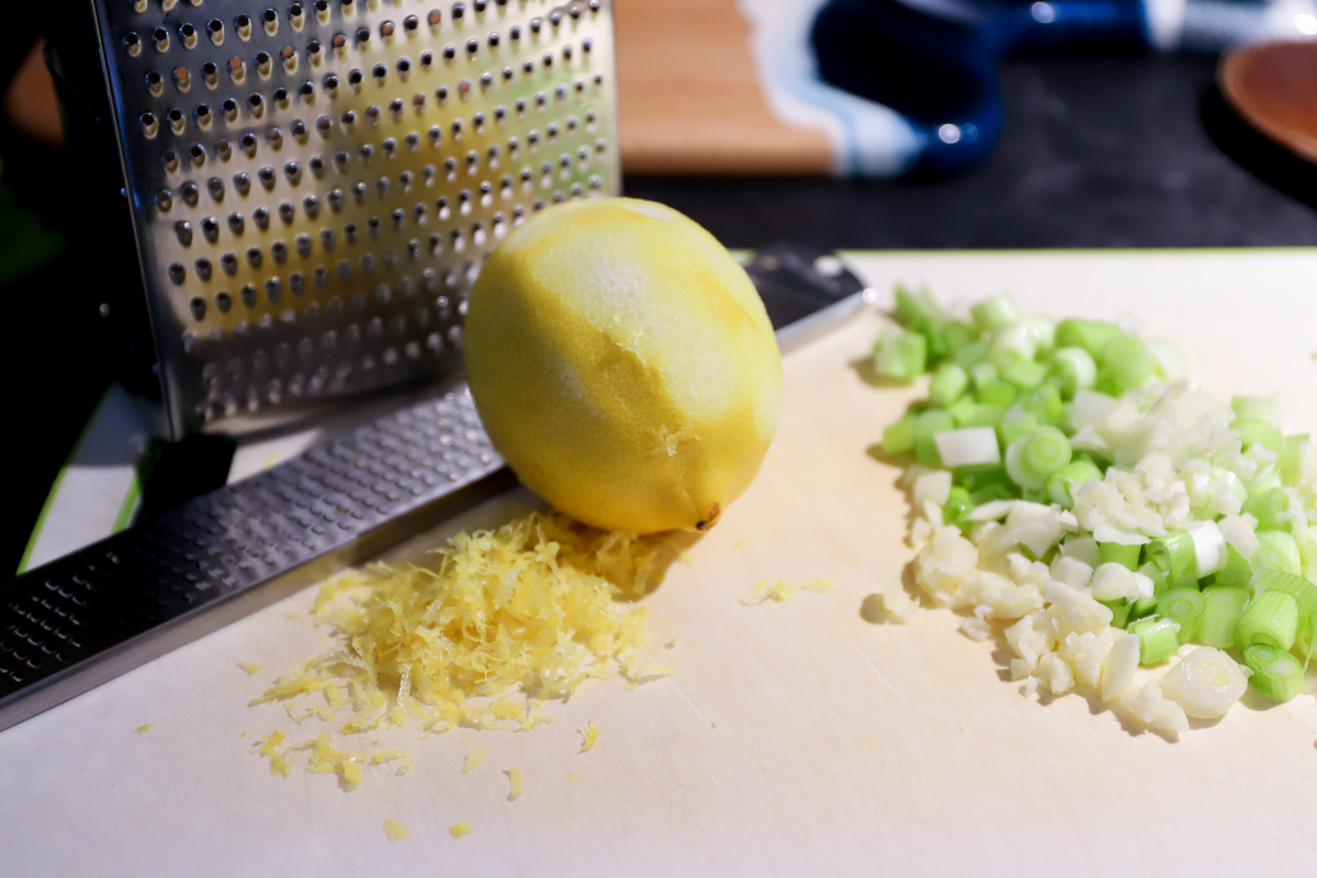 A lemon that has the zest grated off in a pile in front. Green onions are beside the lemon.