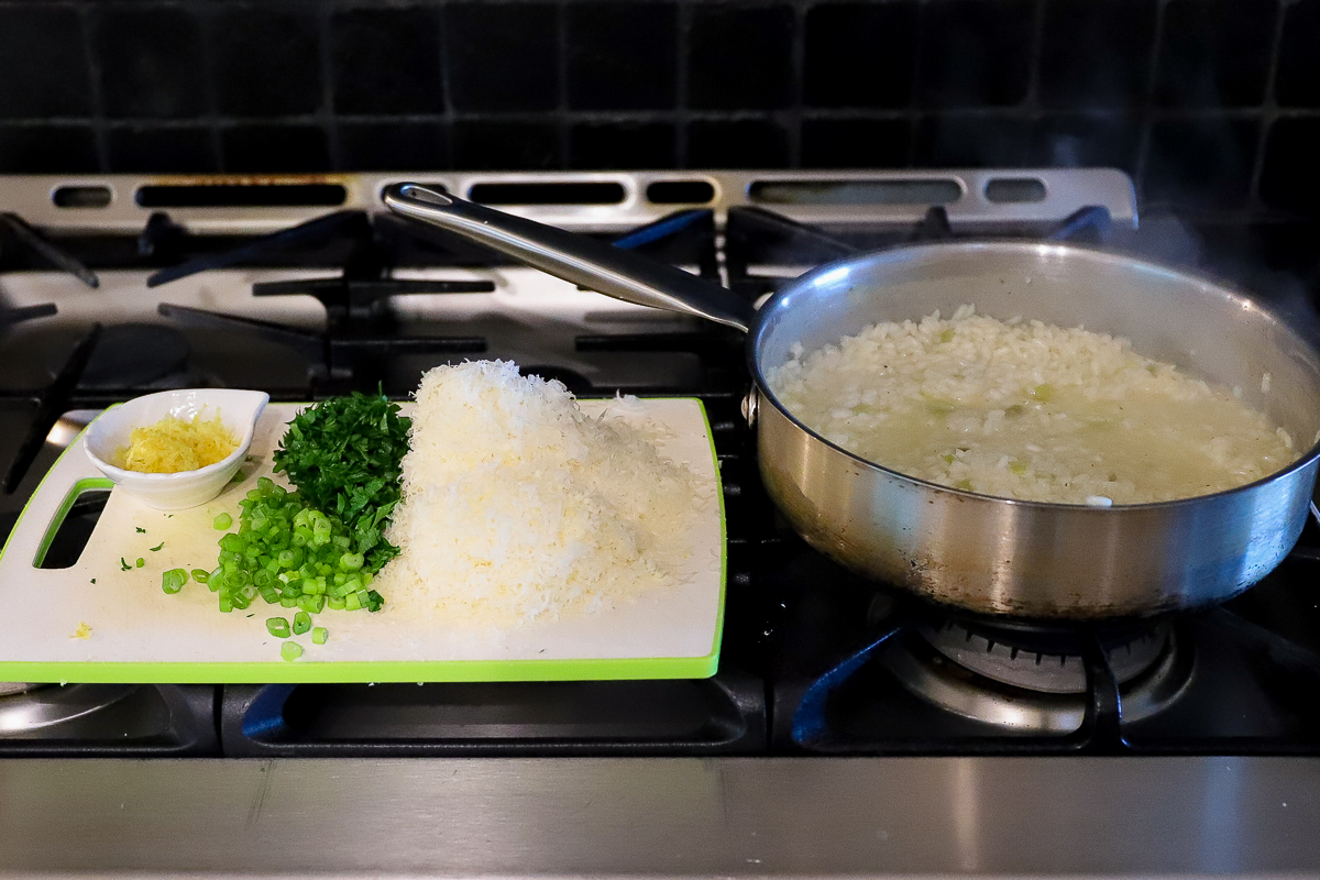 Risotto simmering in saucepan on stove. Cutting board with grated parmesan, parsley, green onion, lemon and lemon juice right beside.