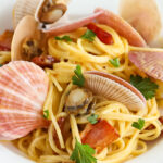 Close up of pink scallops in shell with spaghetti noodles glazed with the sauce and large pieces of bacon.