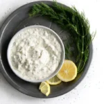 Bowl of tartar sauce with dill fronds and lemon around it.