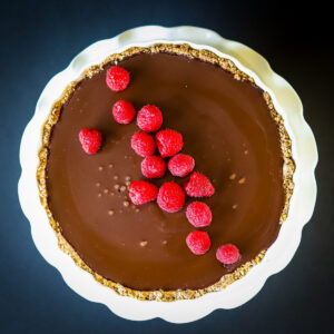 A hazelnut crusted chocolate tart on a fluted white cake plate. There are a few small bubbles in the chocolate that have set and it is garnished with fresh raspberries in a line diagonally across the tart.