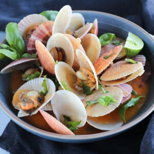 Grey bowl with swimming scallops in shell with red curry broth. Garnished with lime, basil and cilantro.