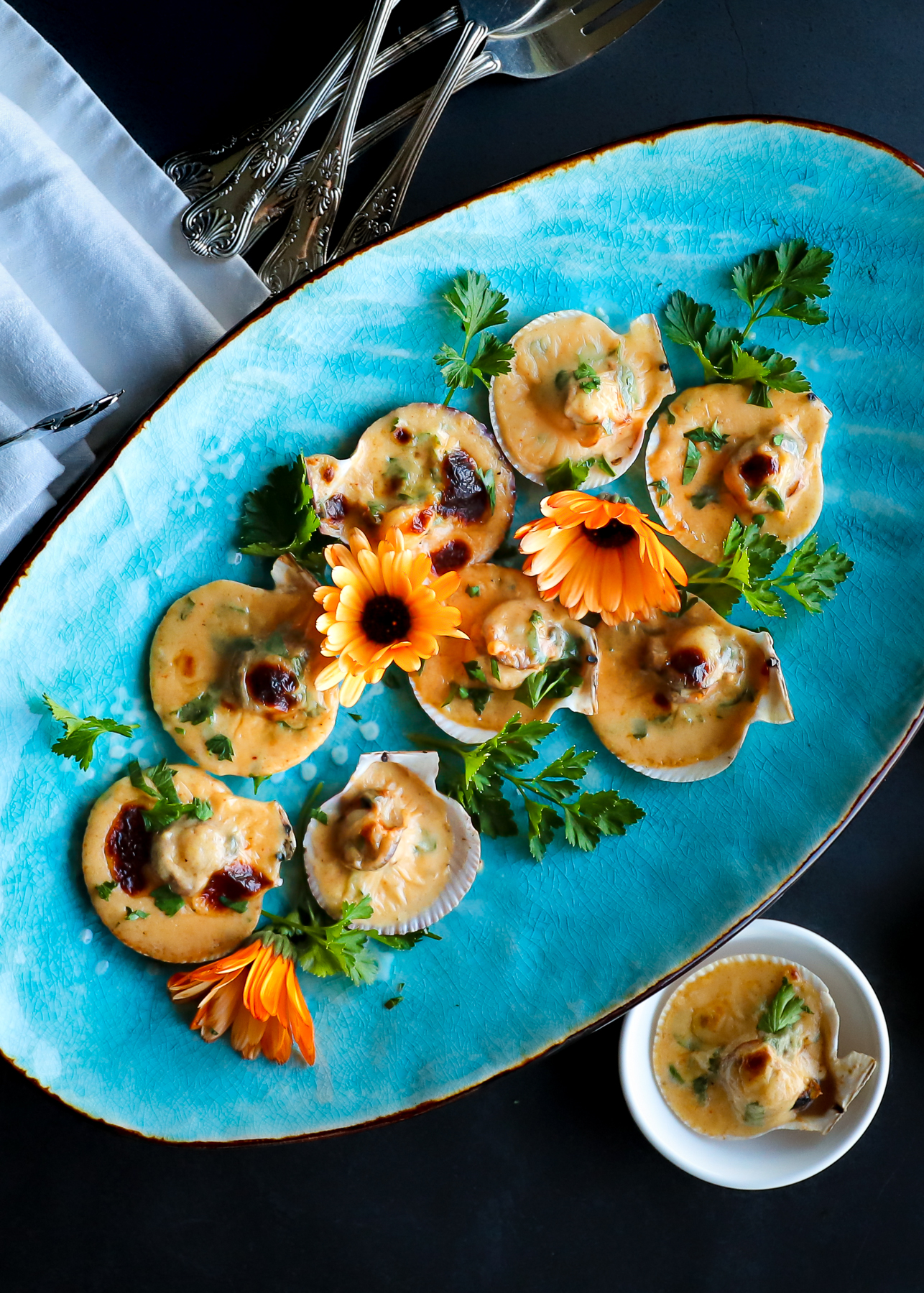 Scallops broiled in their shell arranged on a bright blue plate. Garnished with edible calendula flowers, the plate sits on a black table with forks and white napkins to the left.