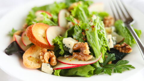 Sliced apples, walnuts, crumbled blue cheese and greens in a white bowl.