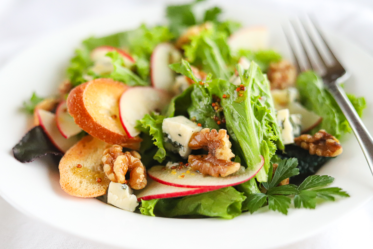 Sliced apples, walnuts, crumbled blue cheese and greens in a white bowl.