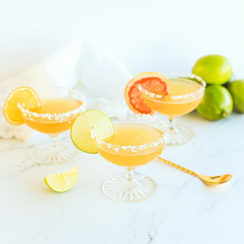 Three margaritas in coupe glasses with salted rims. There is a gold cocktail stirring spoon laying down and a pile of limes in the background. Each drink is garnished with a slice of citrus, lime, grapefruit and orange.
