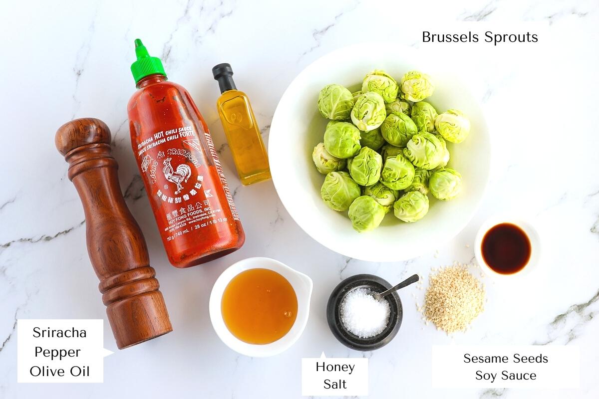 Over head shot with labelled ingredients for the recipe including Brussels sprouts, salt, pepper, sriracha, honey, soy sauce, sesame seeds and olive oil.