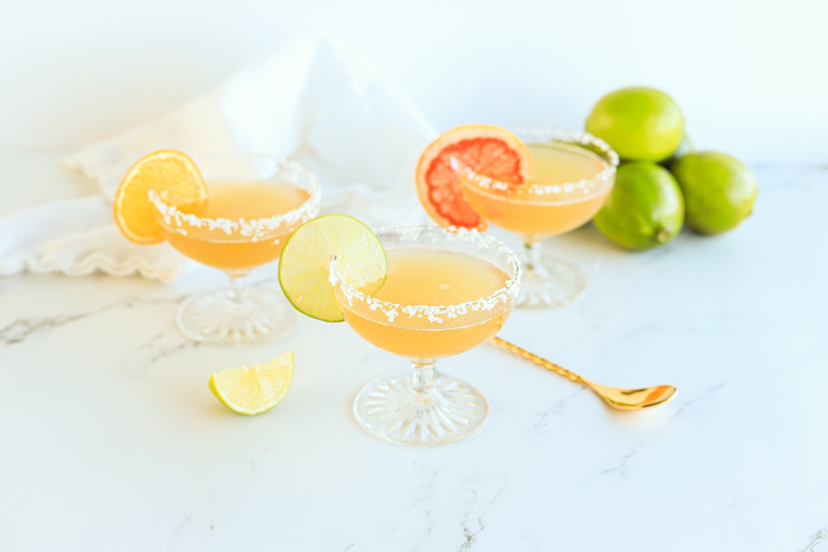 Three margaritas in coupe glasses with salted rims. There is a gold cocktail stirring spoon laying down and a pile of limes in the background. Each drink is garnished with a slice of citrus, lime, grapefruit and orange.