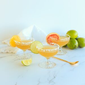 Three margaritas in margarita glasses with salted rims and citrus wheels garnishing them. They are on a white marble countertop with white napkin and gold cocktail spoon.