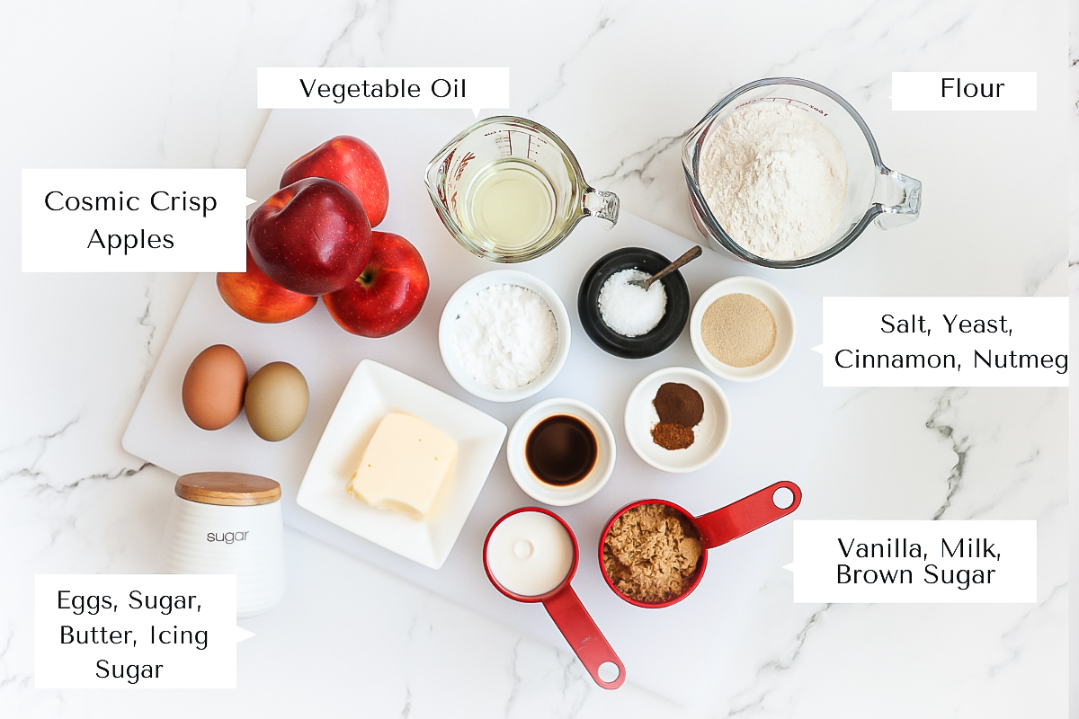 Labelled image of ingredients in an overhead shot. Shown are apples, eggs, sugar, butter, oil, flour, salt, icing sugar, vanilla, brown sugar, milk, cinnamon and nutmeg.