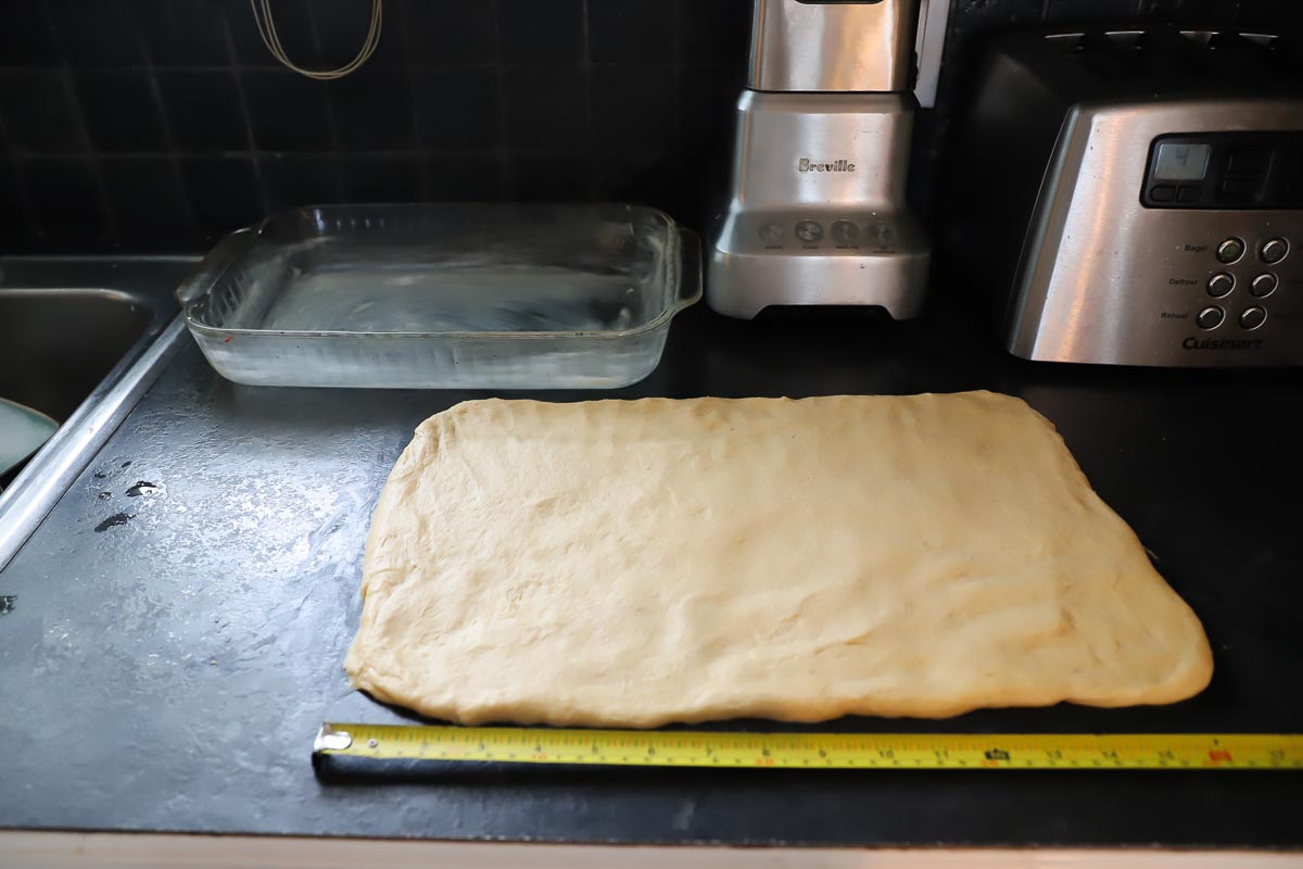 Dough is rolled out on a black countertop with a measuring tape in front. It is in a 12 inch by 16 inch rectangle.