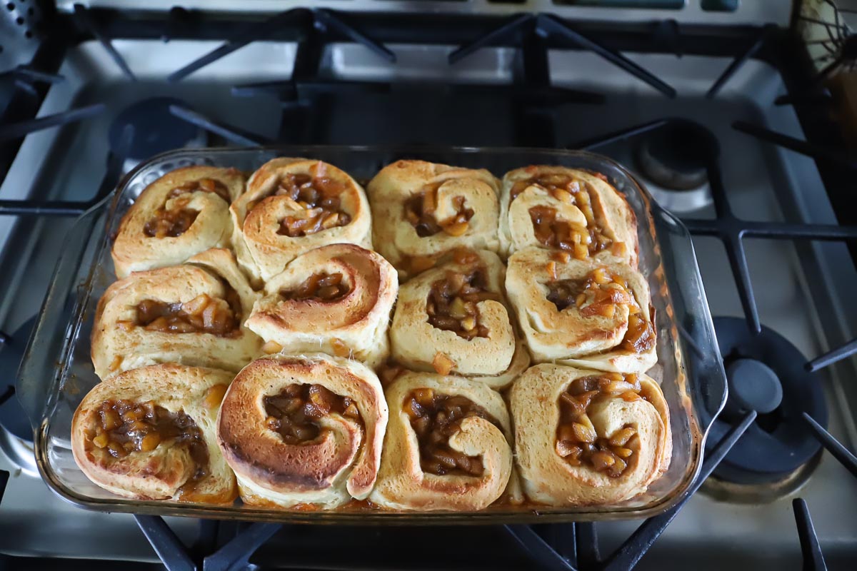 Cinnamon buns are finished baking and have light brown edges. They are sitting on top of the stove in the glass pan cooling.