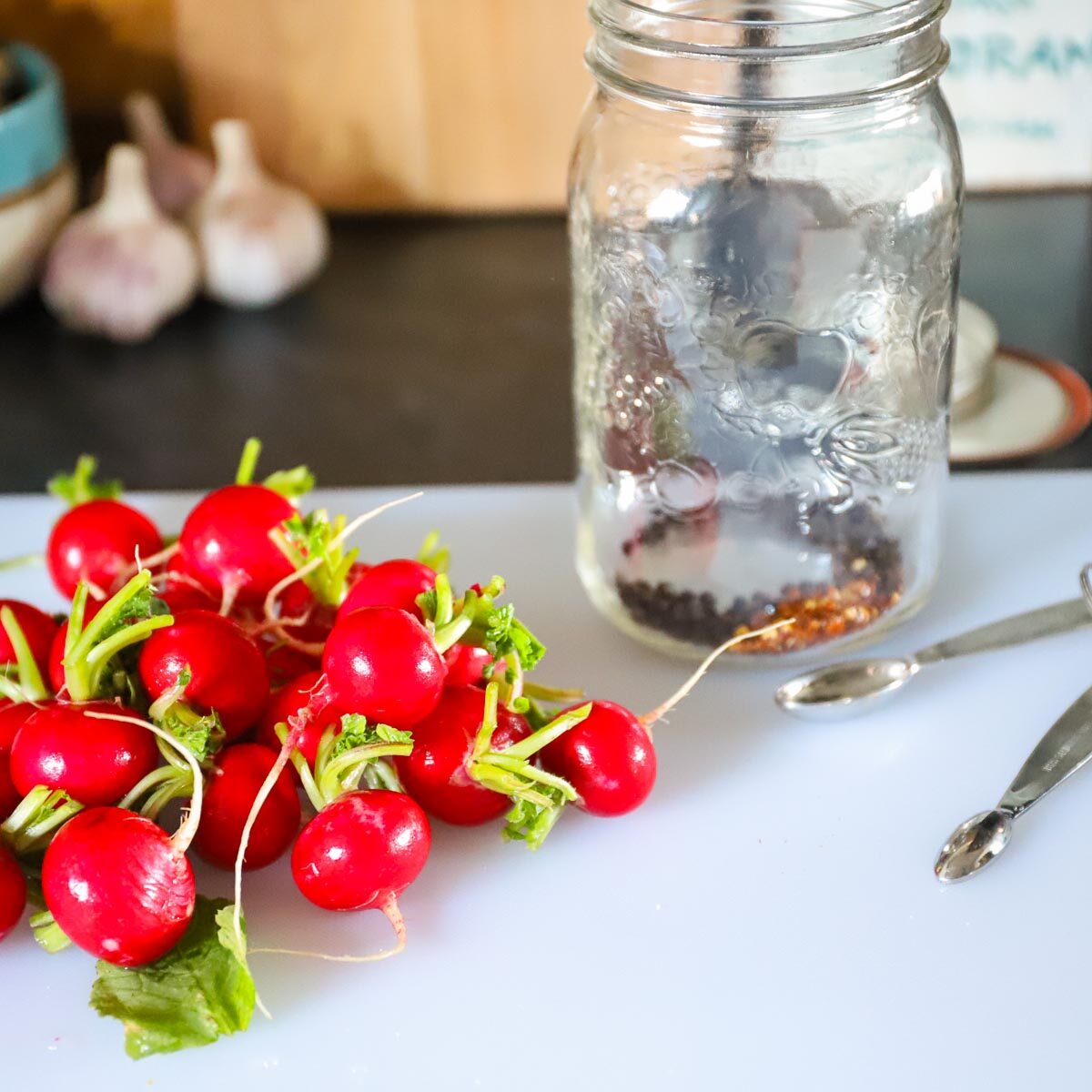 Wet washed radishes on a cutting board beside a wide mouth quart mason jar that has peppercorns and chili flakes in it.