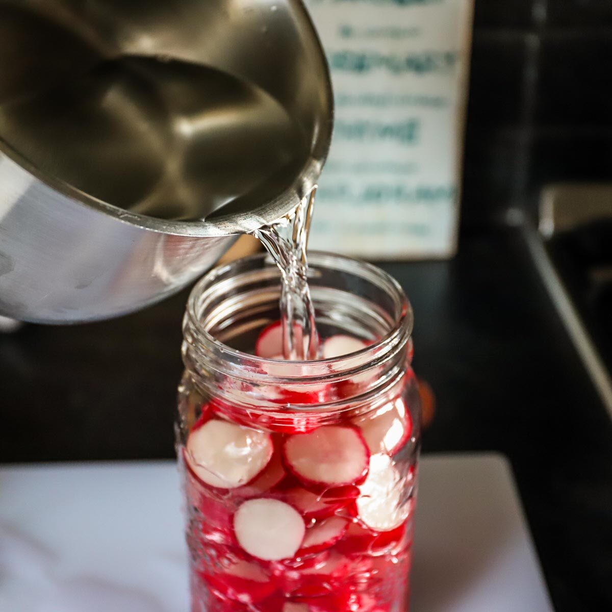 The clear salt water brine is being poured over the radishes in the mason jar.