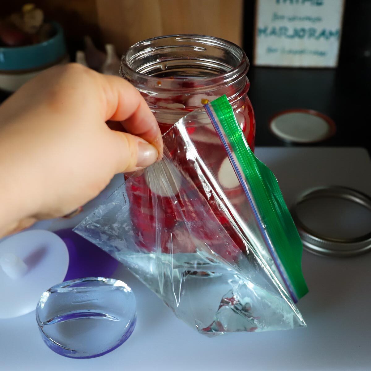 A small, green topped clear zip top baggie is being held up to show the water in the bag that is a makeshift fermenting weight. The jar of radishes is behind.