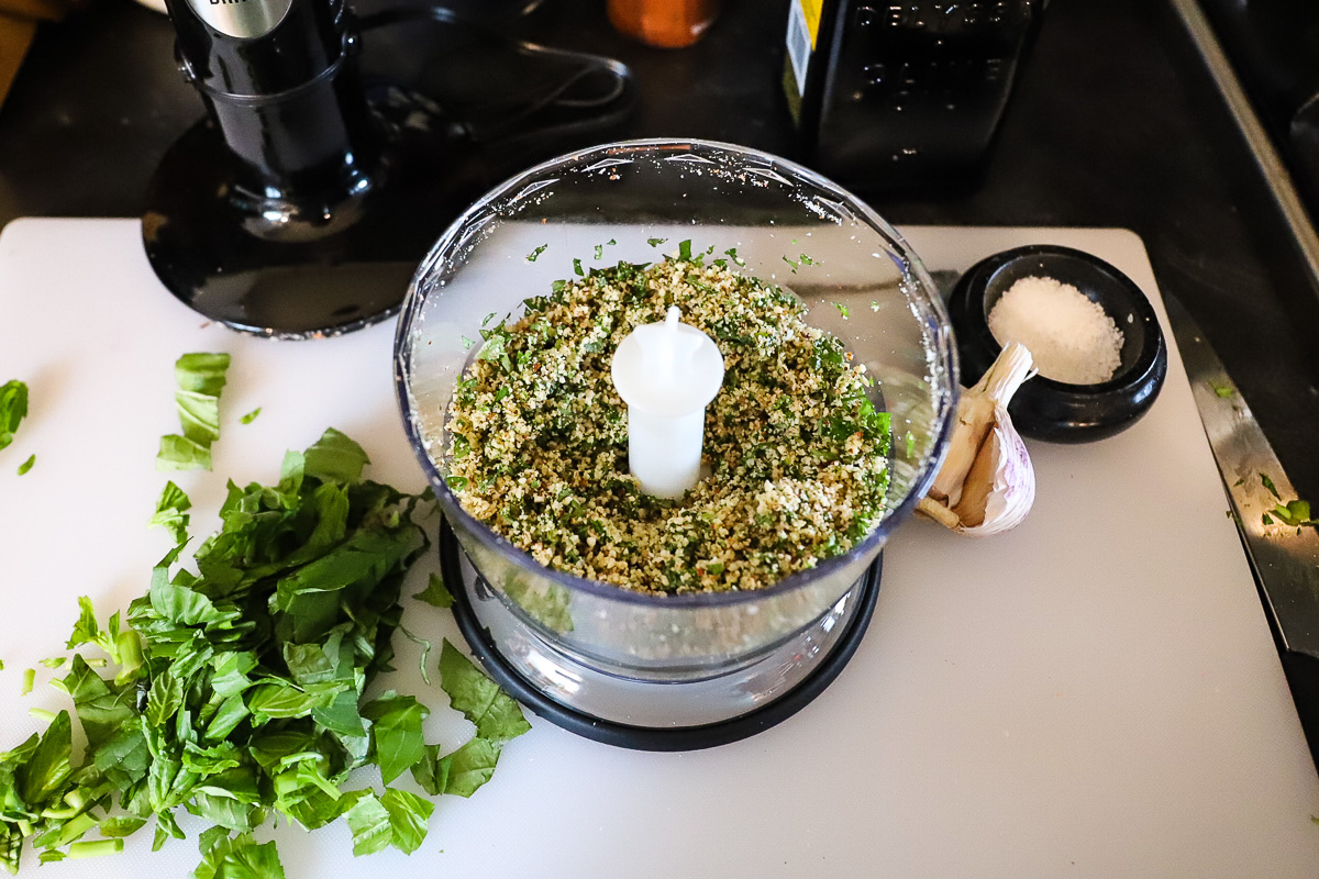 The bowl of the food processor is open, showing the finely chopped almonds and half the basil. The remaining basil is beside the bowl, coarsely chopped.