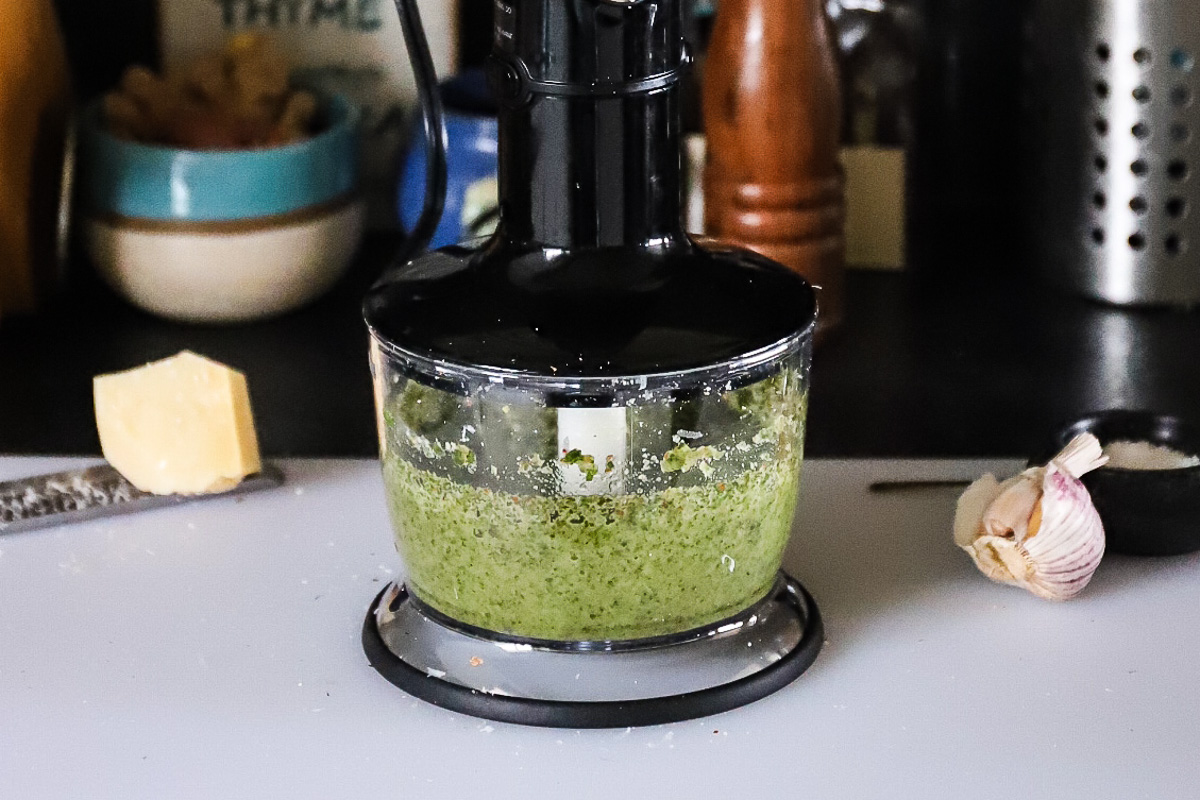 Pesto in the food processor is pale green with very small brown flecks in it. The machine is sitting on a white cutting board with parmesan cheese and garlic nearby.