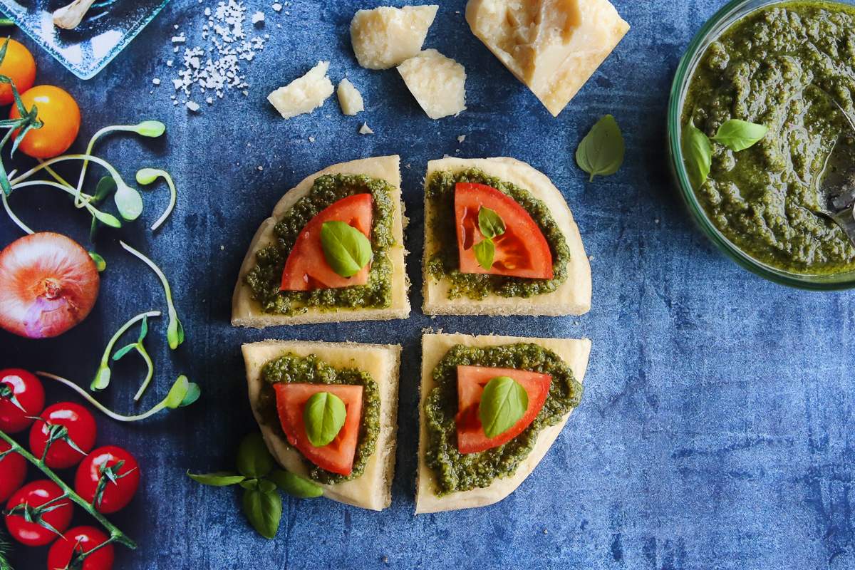 Triangles of bread topped with pesto, tomato and basil leaves. There are other vegetables, parmesan cheese and pesto in a bowl surrounding the bread.