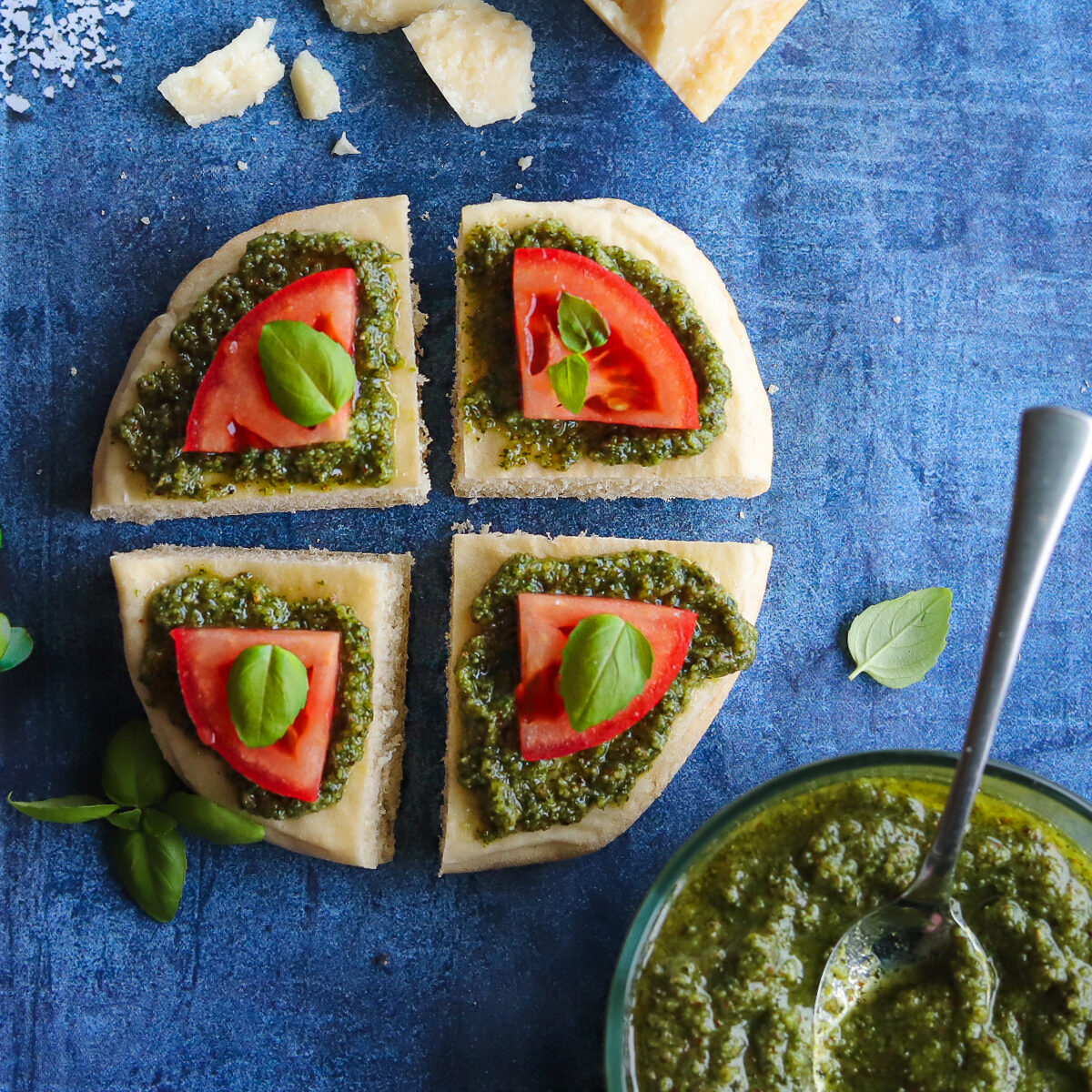 A pita bread cut in four pieces. each piece has a smear of green pesto topped by a quarter slice of tomato and a basil leaf. It is on a blue background with parmesan cheese, pesto and basil leaves around it.
