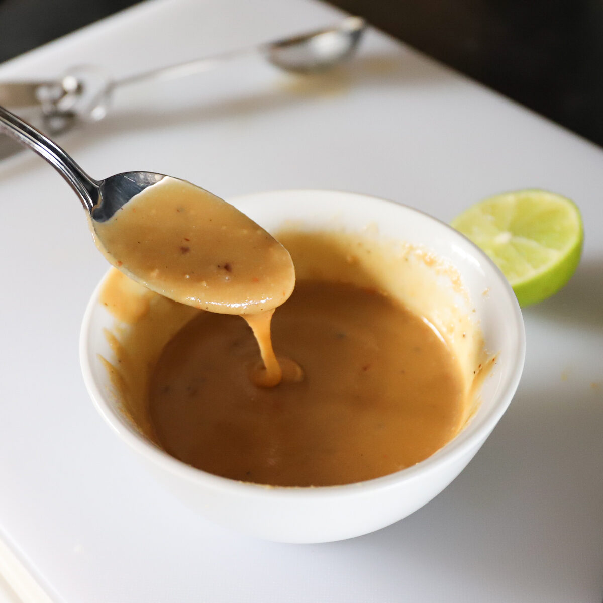 The brown tahini maple dressing in finished in the white bowl. A spoon has a scoop dripping out to show the creamy runny texture.