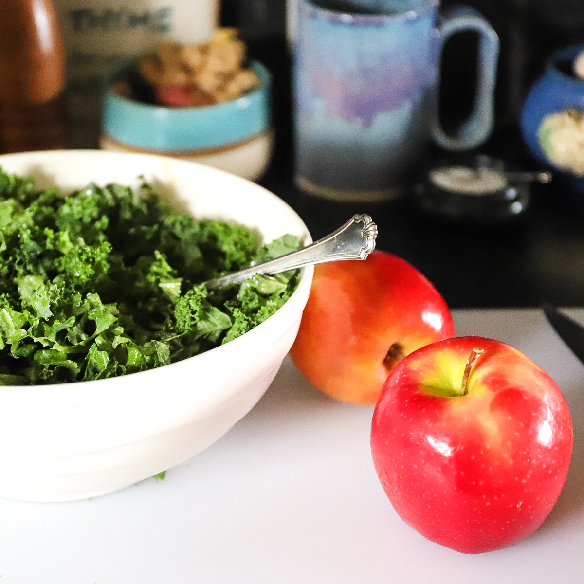 The chopped kale is in a large white bowl. Two whole red apples are sitting beside the bowl..